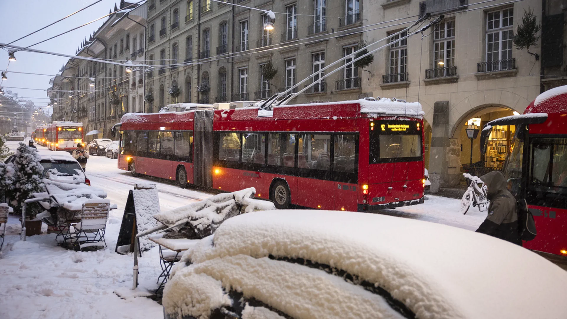 Schnee auf Autos und Tischen von Restaurants. Auf der Strasse stehen mehrere Busse.