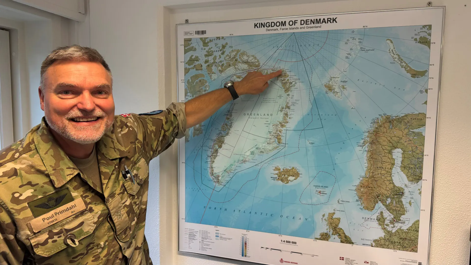 Ein Mann mit hellblondem Haar in Tarnfarben-Uniform zeigt auf eine Karte, darauf die Insel Grönland.