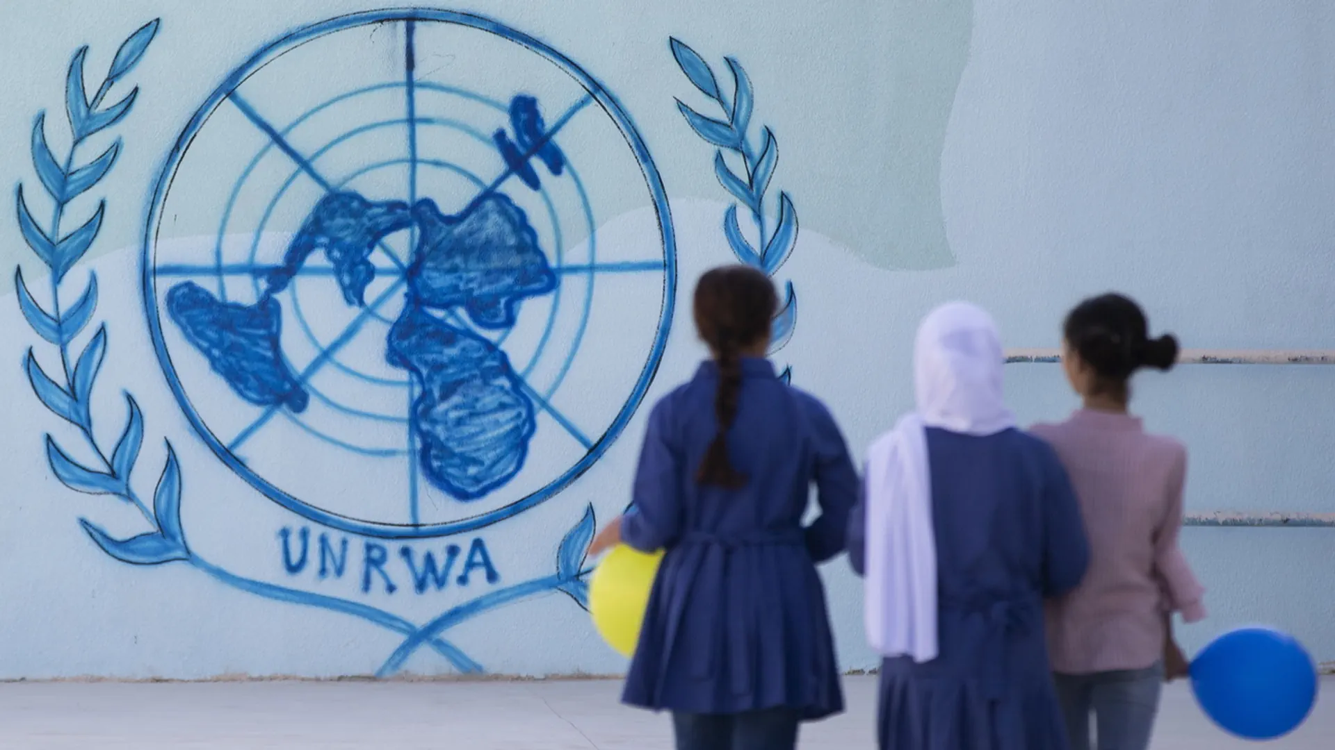 An einer Wand ist das Logo der UNRWA aufgemalt. Im Vordergrund sieht man von hinten schemenhafte Gestalten.