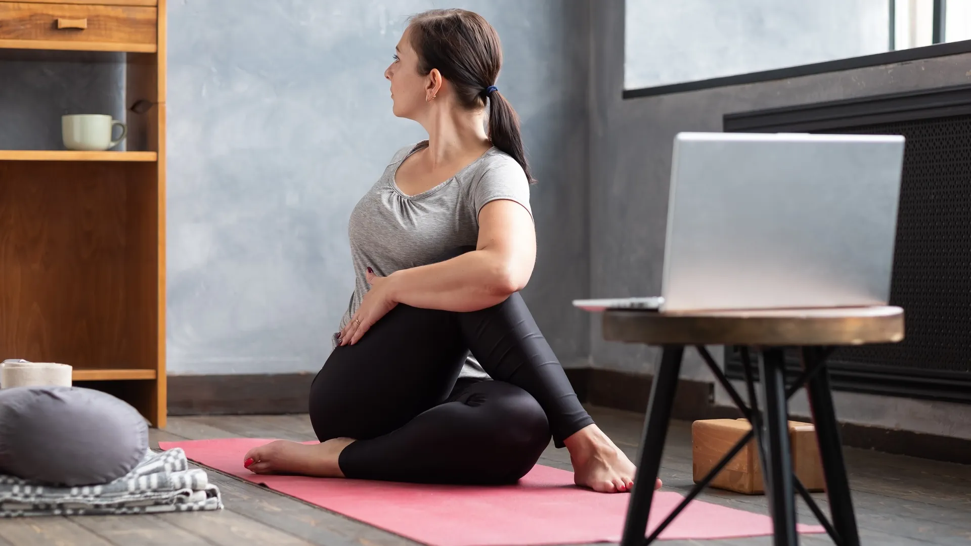 Frau macht auf einer Matte sitzend eine Yogabewegung vor dem Laptop